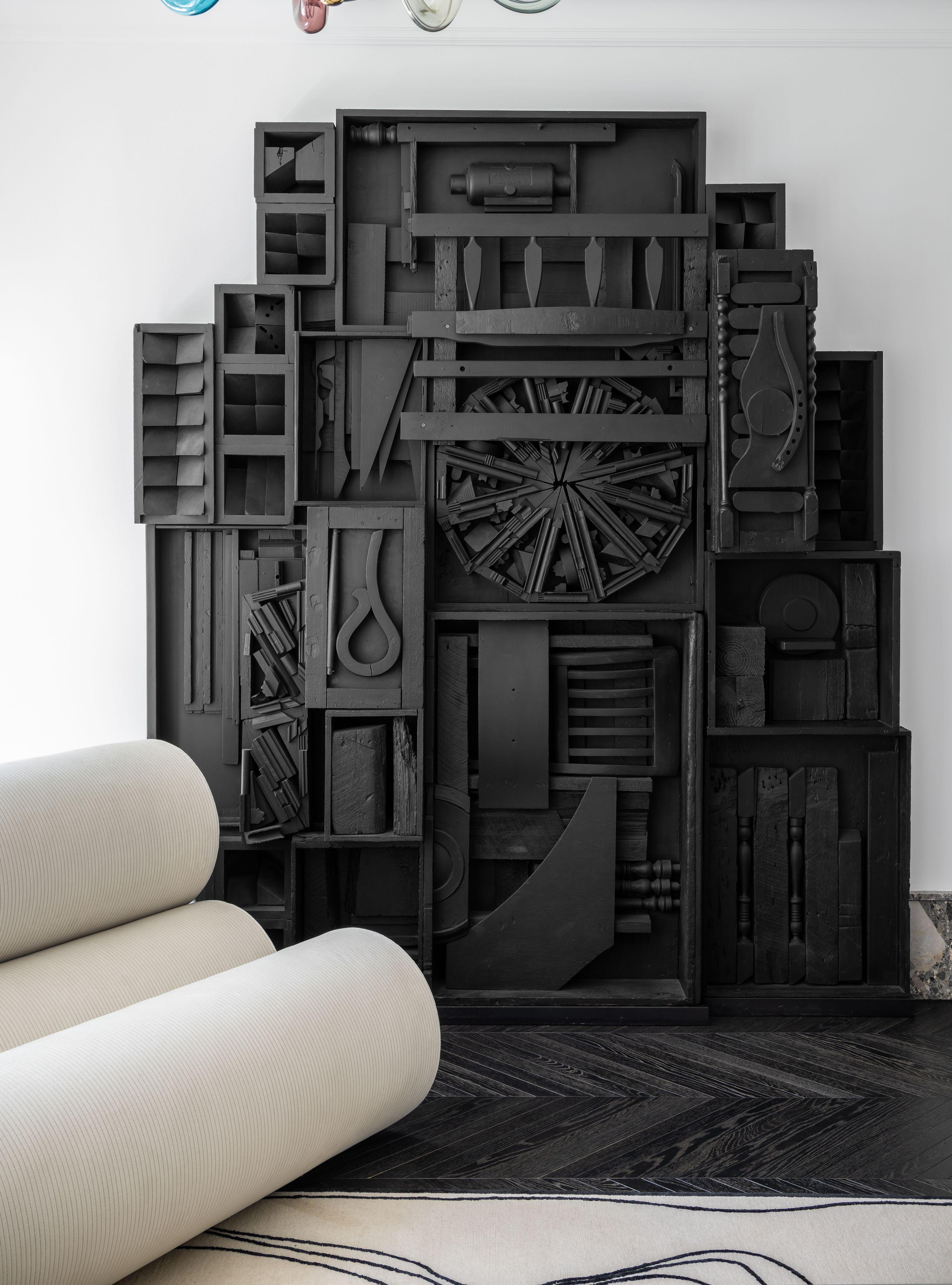 This interior composition shows a monumental black assemblage wall sculpture, likely constructed from found wooden forms — drawers, beams, tools, and architectural fragments — arranged into a relief recalling Louise Nevelson’s style