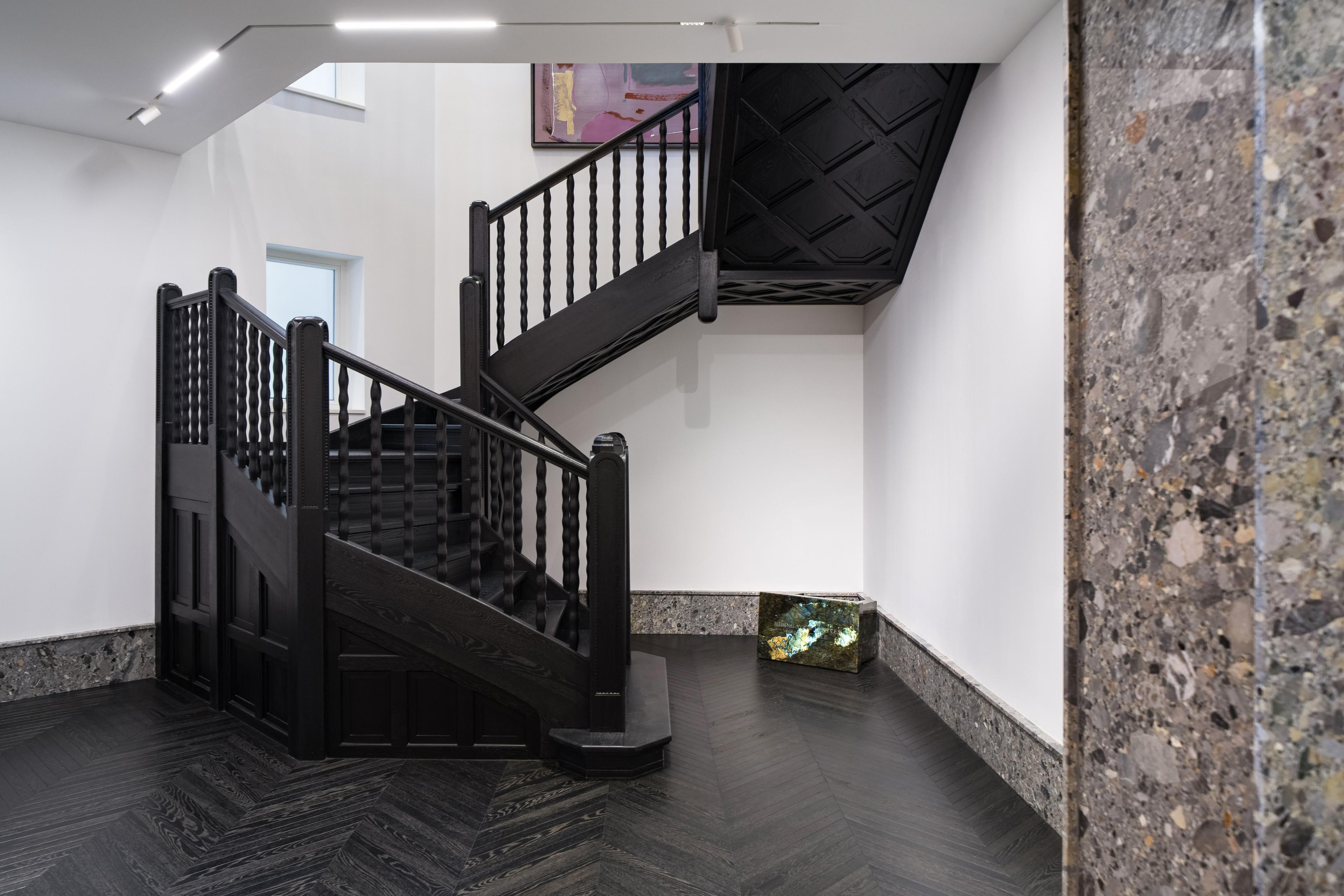 A dark wood staircase with geometric detailing ascends in a minimalist gallery space featuring terrazzo accents and a reflective sculptural installation in the corner.
