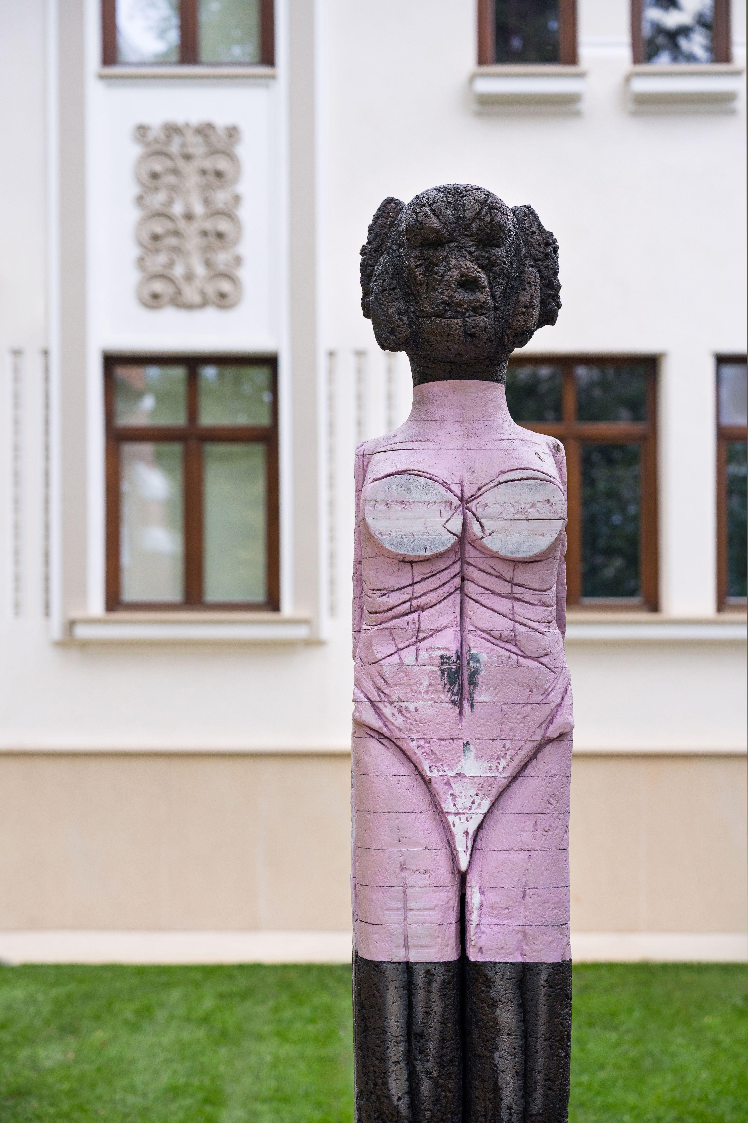 A stylized, upright female form — carved wood with a heavily textured, charred or earthen surface at the head and lower half, and painted in soft pink hues across the torso.